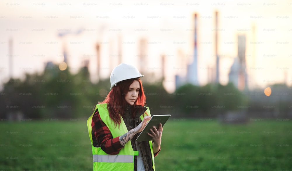 Young woman engineer with hard hat and tablet standing outdoors by oil refinery. Copy space.