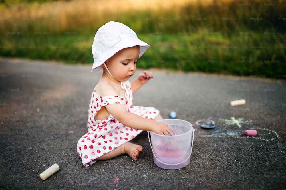 A cute toddler girl outdoors in countryside, chalk drawing on road.