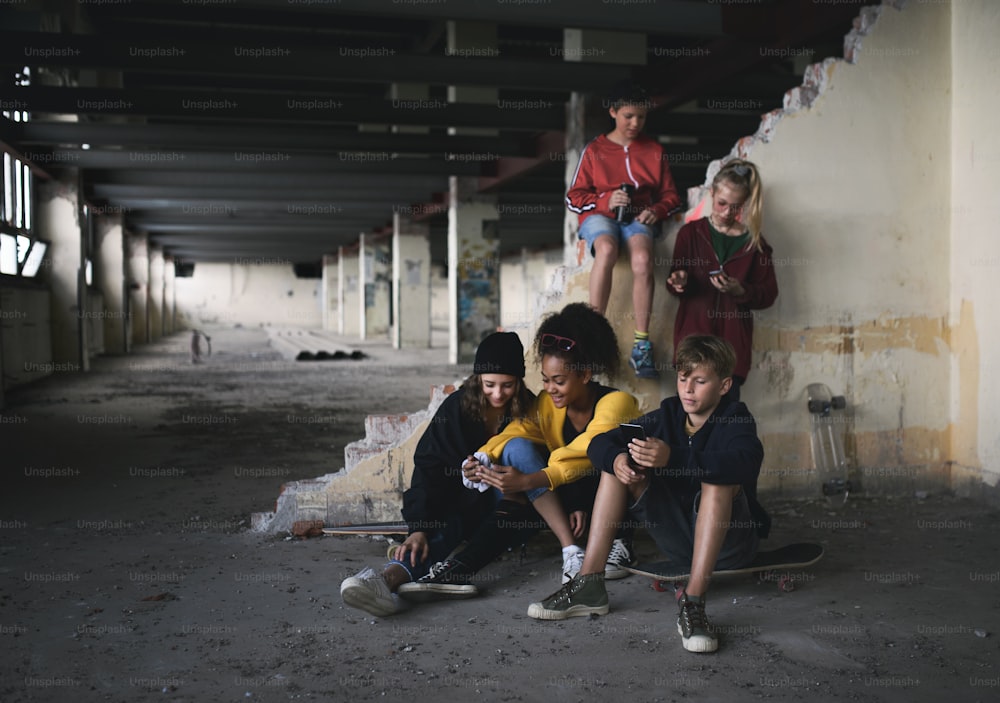 Front view of group of teenagers gang sitting indoors in abandoned building, using smartphones.