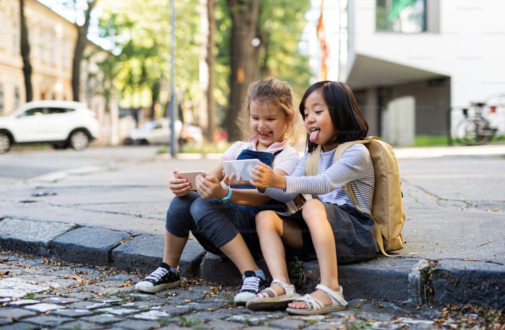 Petites filles avec des smartphones assises à l’extérieur en ville, prenant des selfies.