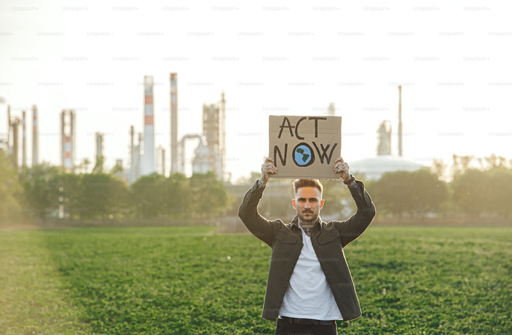 Retrato de un joven activista con un cartel de pie al aire libre junto a una refinería de petróleo, protestando.