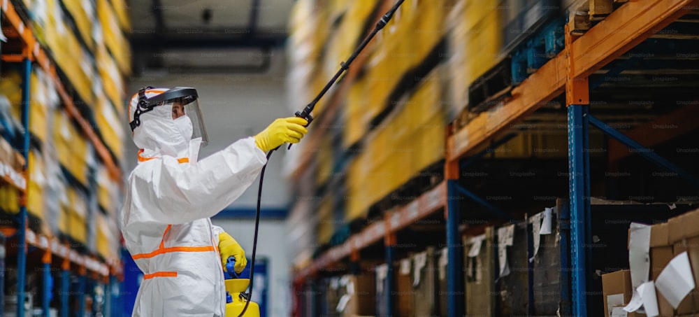 Side view portrait of man worker with protective mask and suit disinfecting industrial factory with spray gun.