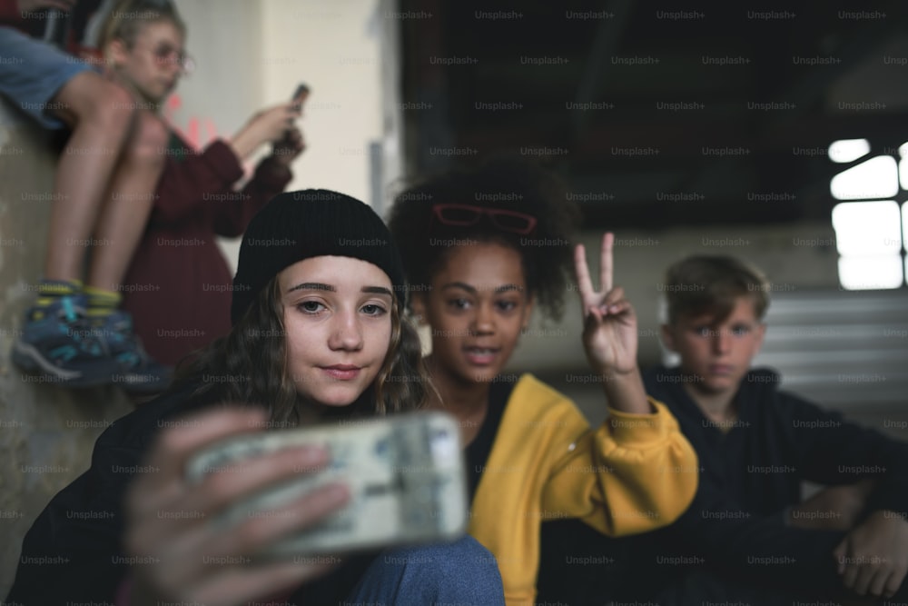 Front view of group of teenagers gang sitting indoors in abandoned building, taking selfie with smartphone.