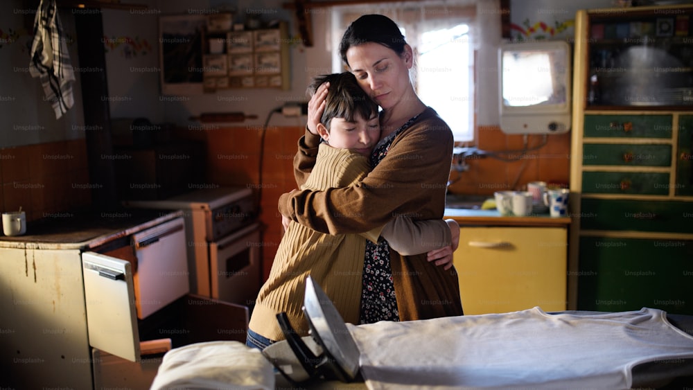 Ein Porträt einer traurigen armen reifen Mutter, die ihre kleine Tochter zu Hause umarmt, Armutskonzept.