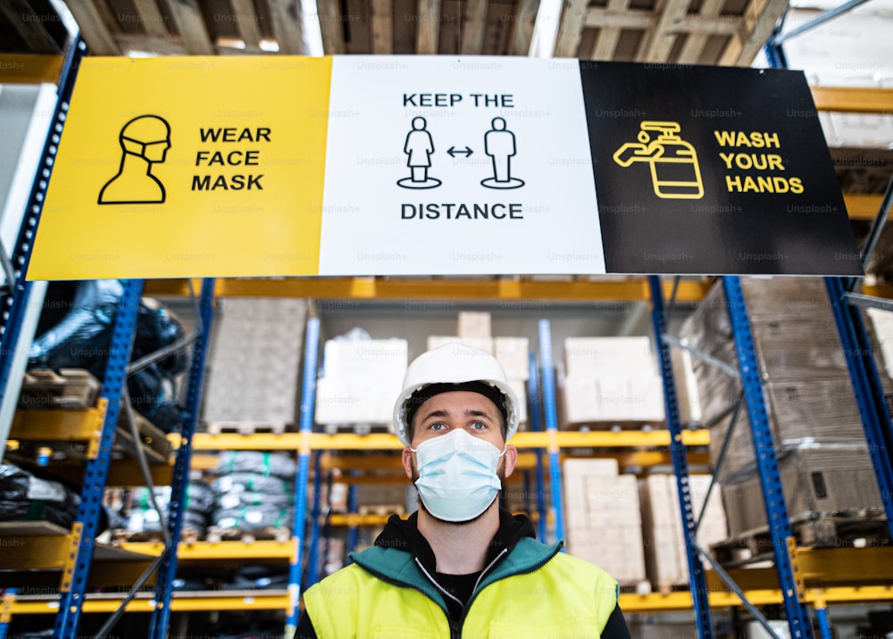 Portrait of young worker with face mask standing indoors in warehouse, coronavirus concept.