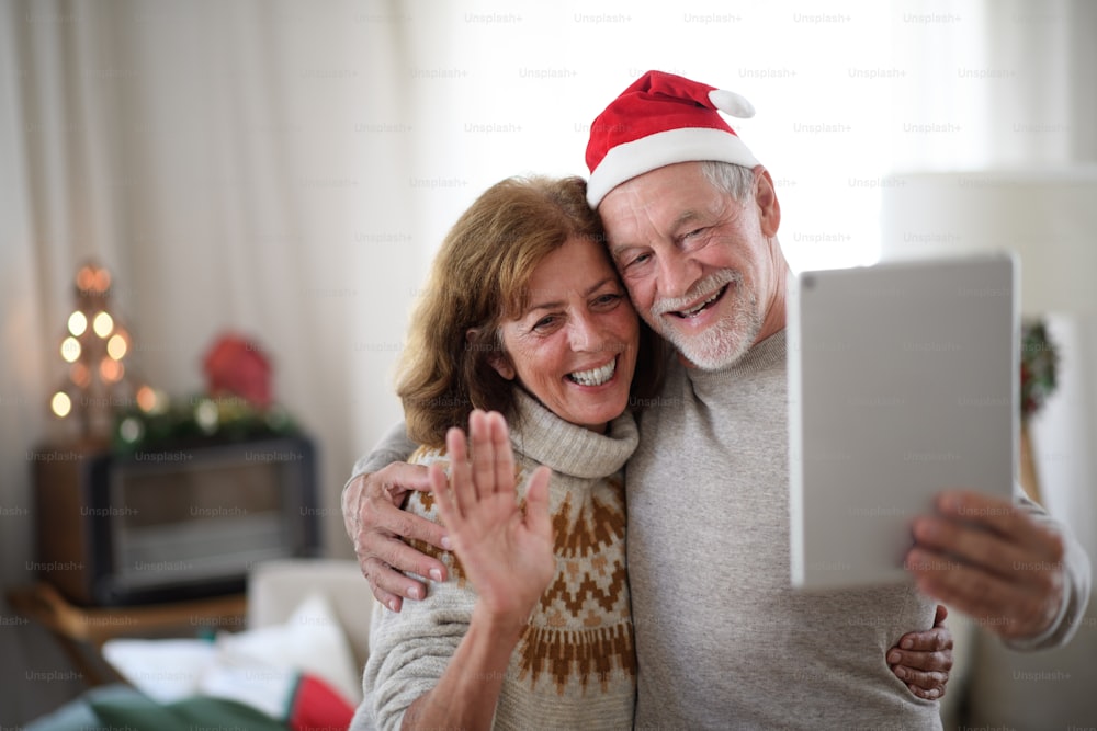 Front view of happy senior couple with face masks indoors at home at Christmas, taking selfie with tablet.