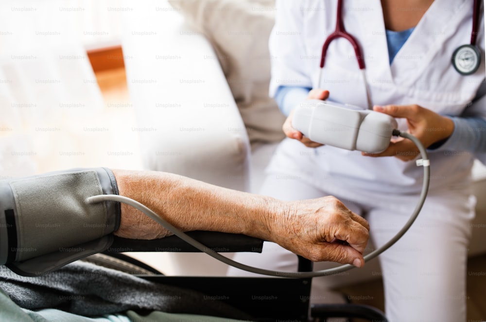 Visitador de salud irreconocible y una mujer mayor durante la visita domiciliaria. Una enfermera revisando la presión arterial de una mujer en silla de ruedas.