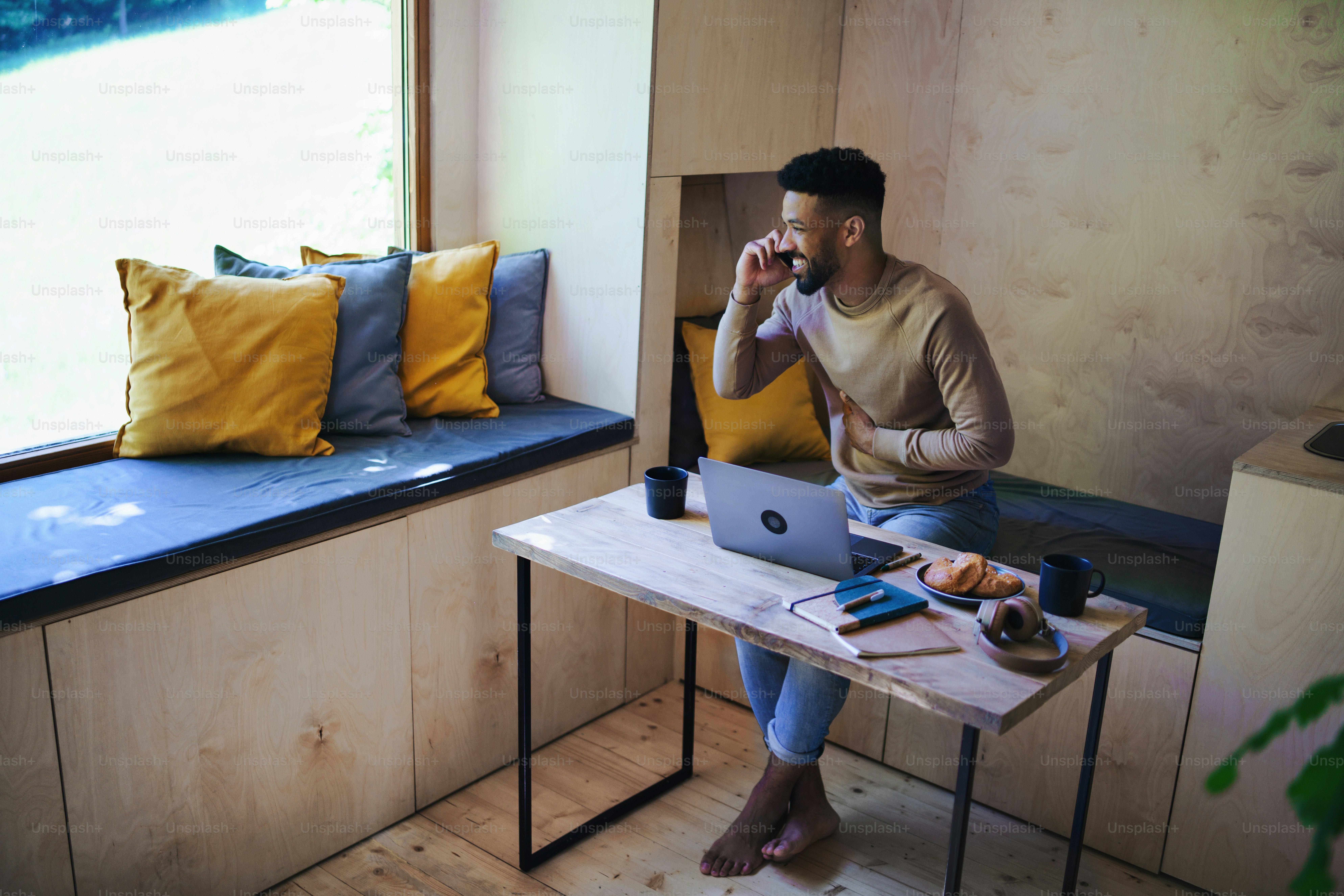 Ein glücklicher junger Mann mit Laptop und Smartphone sitzt, ruht sich drinnen in einem Baumhaus, Wochenendausflug und Remote-Office-Konzept aus.