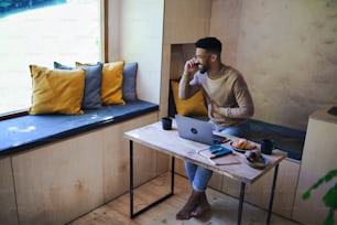 Un jeune homme heureux avec un ordinateur portable et un smartphone assis, se reposant à l’intérieur dans une cabane dans les arbres, un week-end et un concept de bureau à distance.