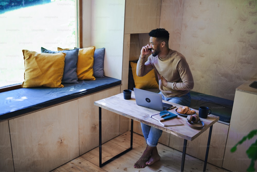 Un joven feliz con una computadora portátil y un teléfono inteligente sentado, descansando en el interior en una casa en el árbol, un fin de semana fuera y un concepto de oficina remota.
