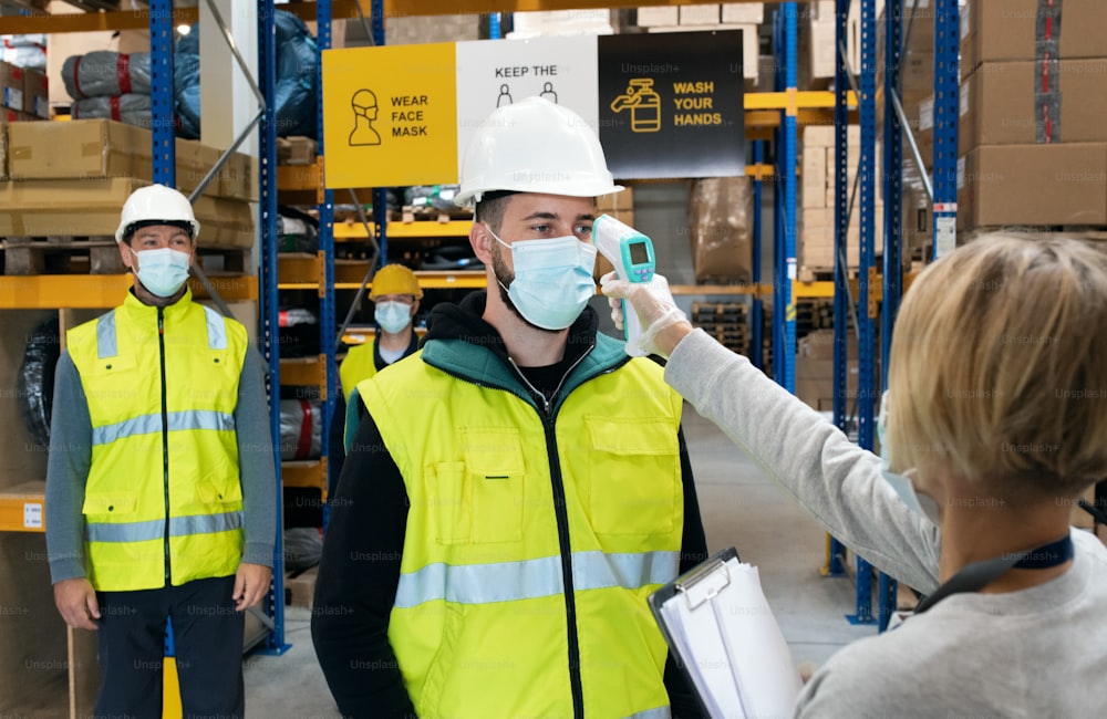 Gruppo di lavoratori con maschera facciale in piedi in magazzino, coronavirus e concetto di misurazione della temperatura.