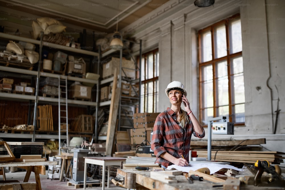 Una ingeniera sosteniendo planos y haciendo llamadas telefónicas en el interior de un taller de carpintería.