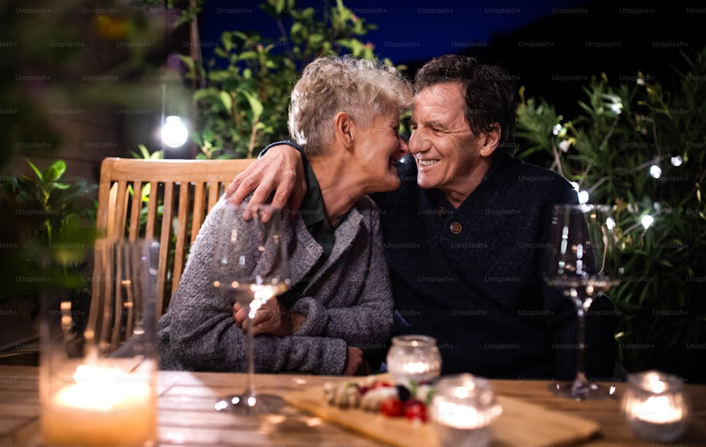 Portrait d’un couple de personnes âgées avec du vin le soir sur la terrasse, s’étreignant.
