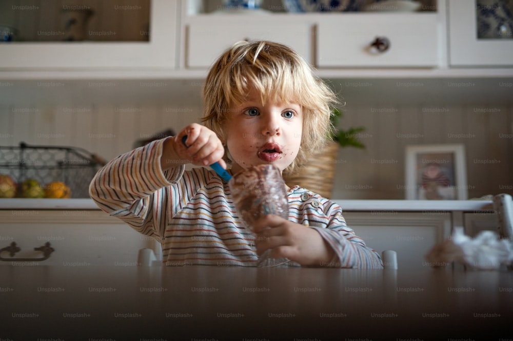Porträt eines kleinen Jungen mit schmutzigem Mund drinnen in der Küche zu Hause, der Pudding isst.