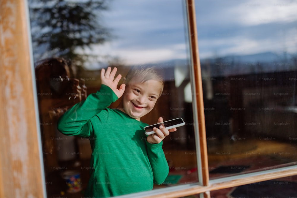Un bambino felice con la sindrome di Down che usa lo smartphone e saluta attraverso la finestra