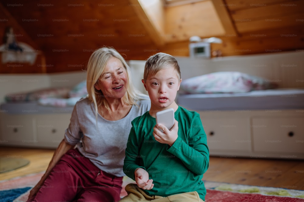 A boy with Down syndrome with his grandmother sitting on floor and using smartphone at home.