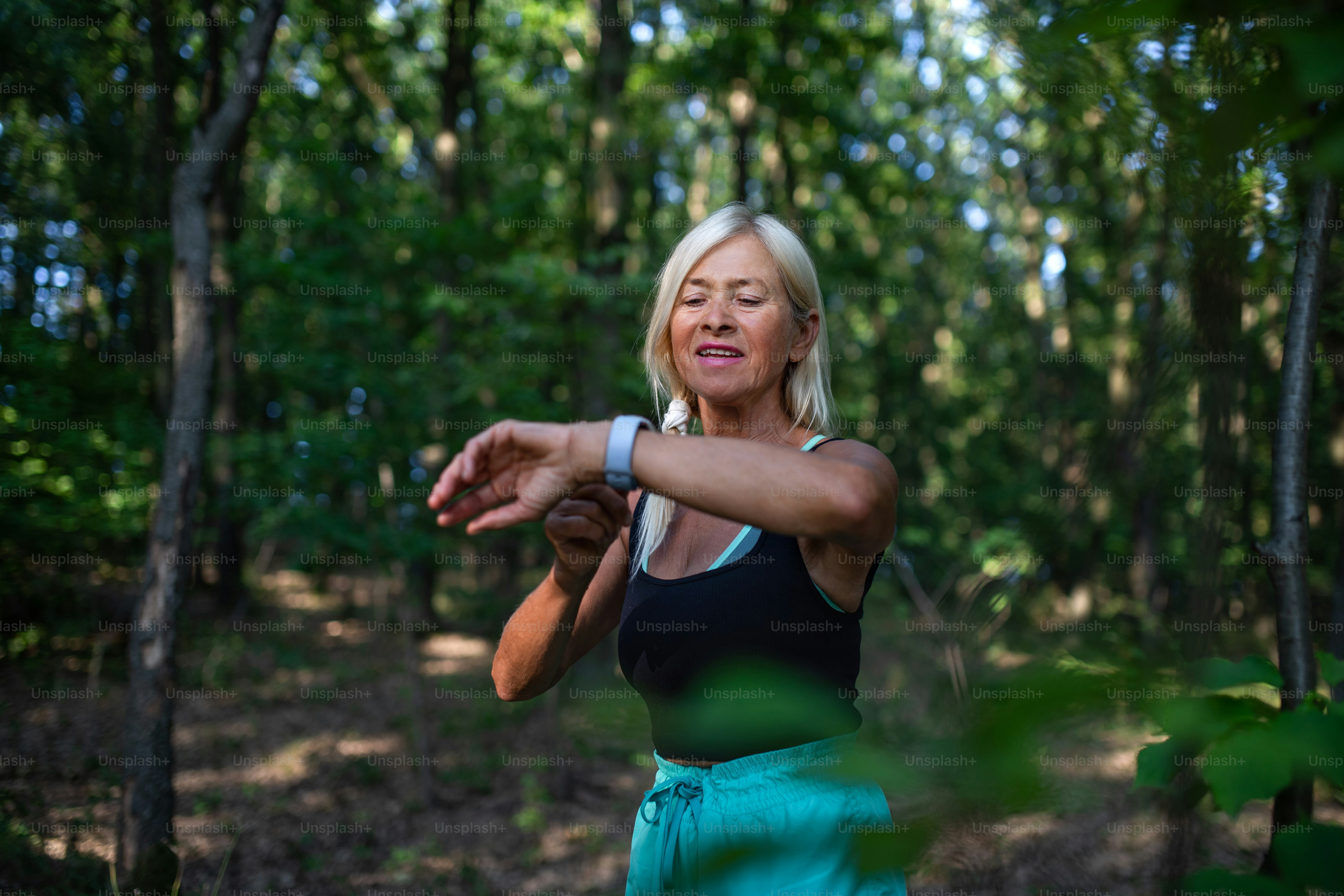 A portrait of active senior woman runner standing outdoors in forest, setting smartwatch.