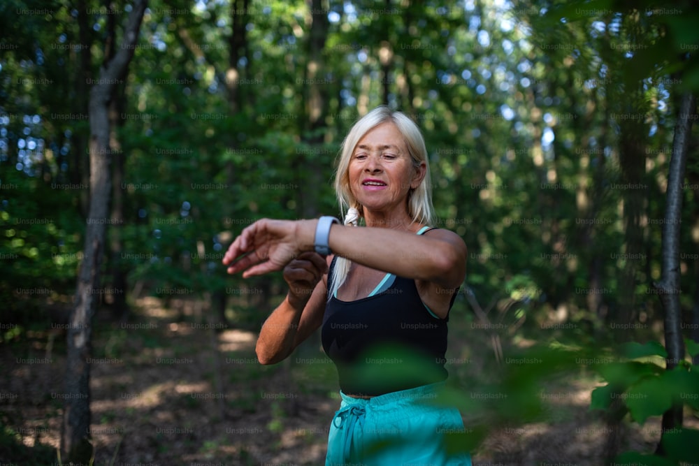 森の屋外に立ち、スマートウォッチをセットするアクティブなシニア女性ランナーのポートレート。