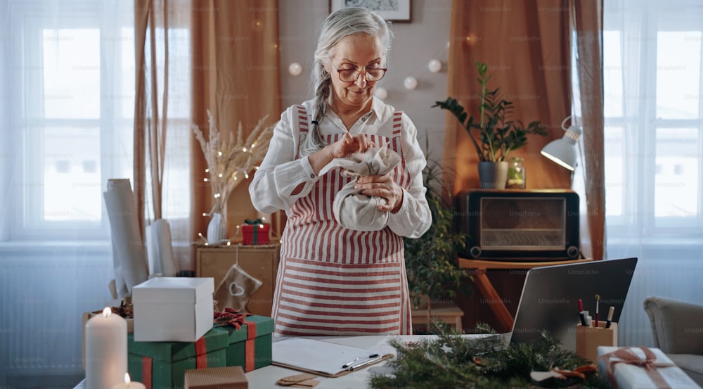 A senior woman packing Christmas present in natural eco materials indoors at home