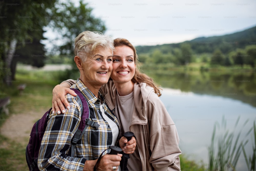 A happy senior mother hiker embracing with adult daughter when looking at lake outdoors in nature