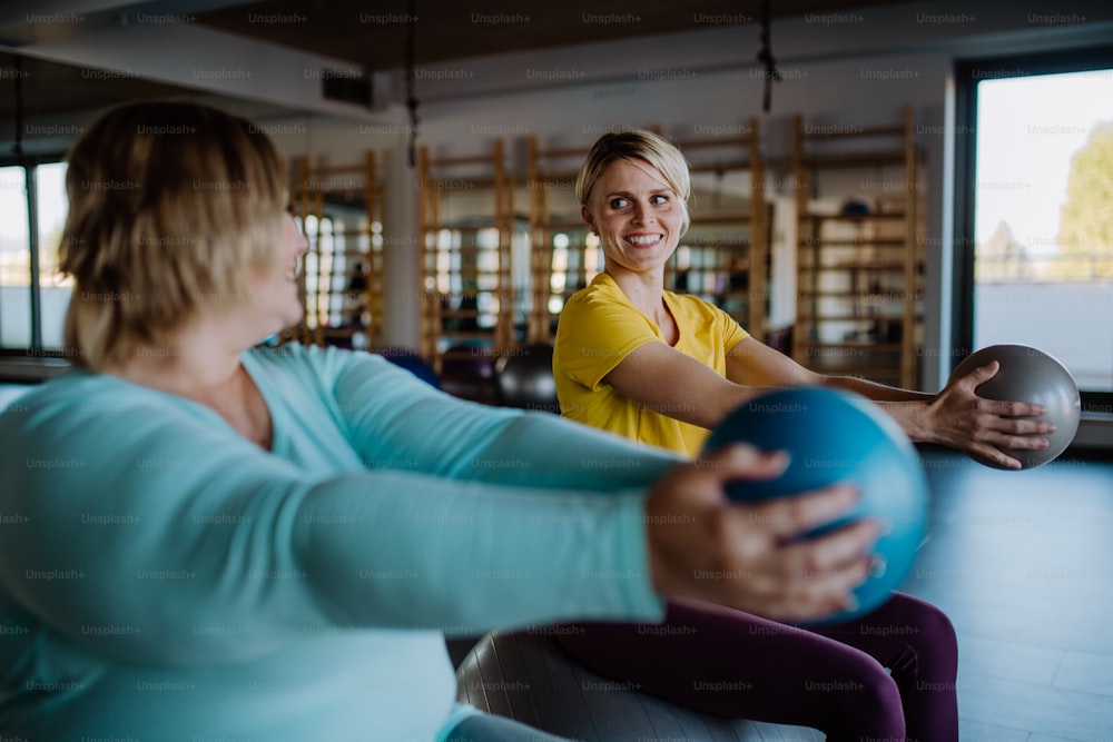 A happy overweight woman sitting on fintess ball and exercising with personal trainer in gym