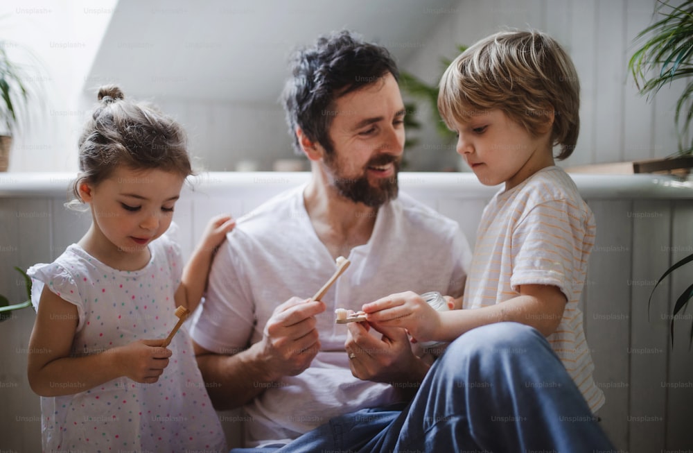 Ein Vater mit zwei kleinen Kindern putzt drinnen zu Hause Zähne, nachhaltiges Lifestyle-Konzept.