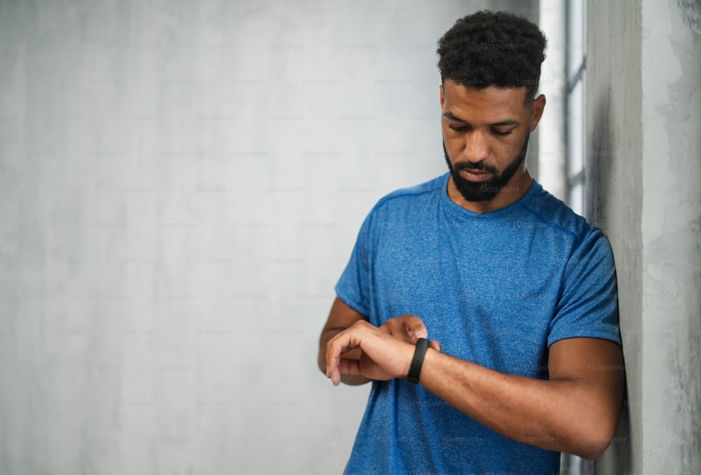 A happy young African American sportsman standing indoors at gym, using smartwatch, copy space.