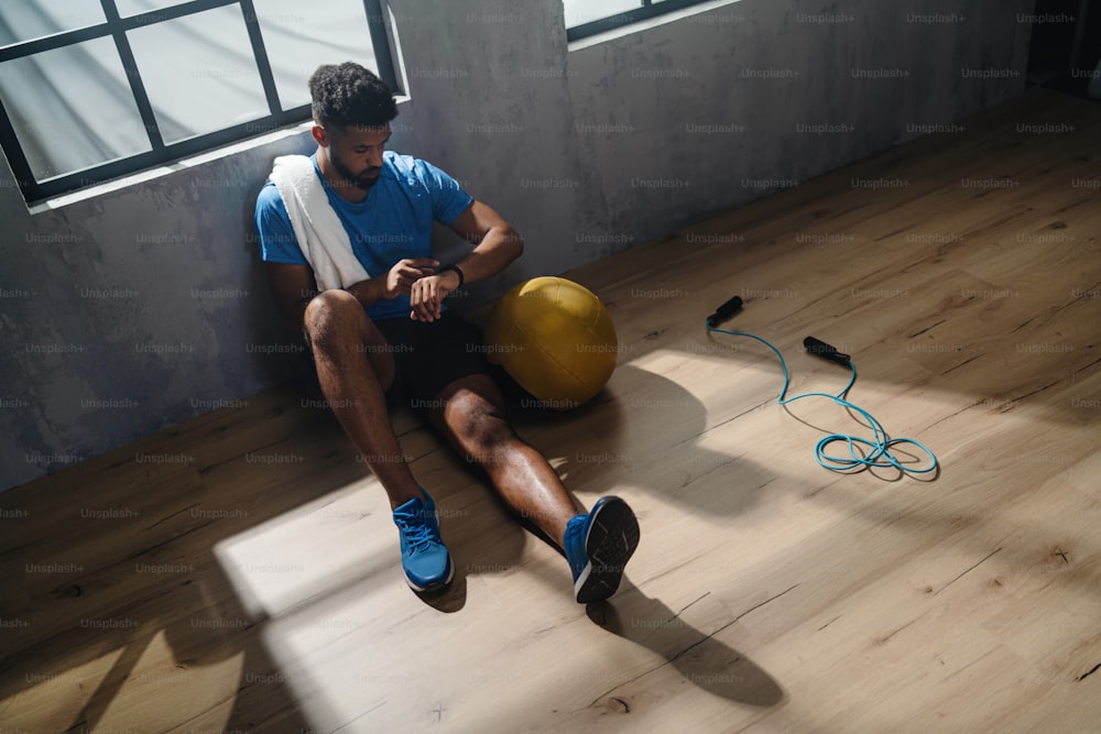 A high angle view of young African American sportsman sitting and using smartwatch indoors in gym.