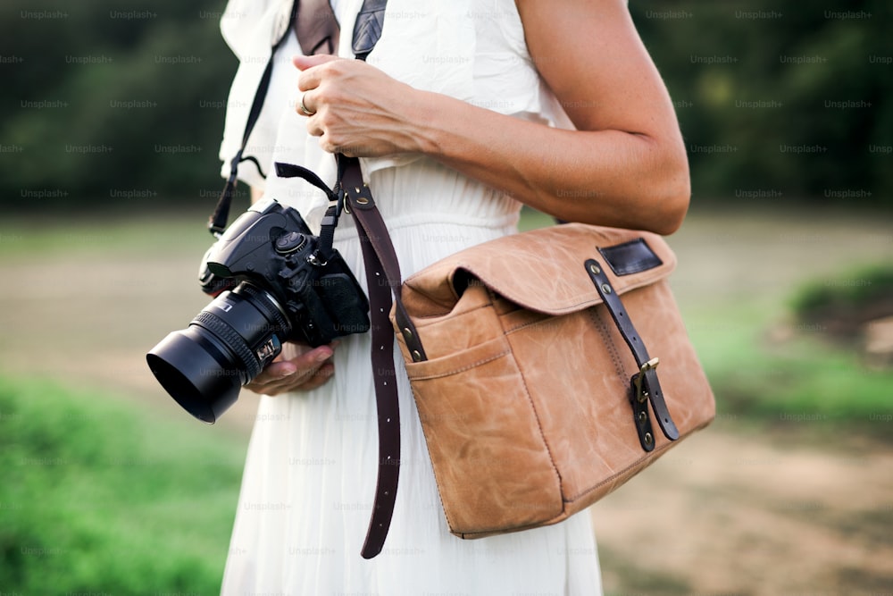 Ein Mittelteil einer nicht wiederzuerkennenden Frau in der Natur, die eine Kamera und eine braune Ledertasche hält.