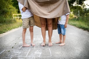 Two small sons playing with cheerful mother on a road in park on a summer day, hiding under her skirt. A midsection.