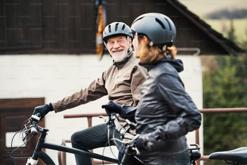 An active senior couple with helmets and electrobikes standing outdoors in front of a house.