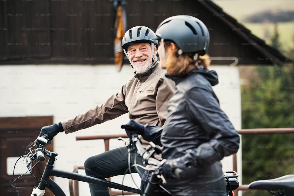 An active senior couple with helmets and electrobikes standing outdoors in front of a house.