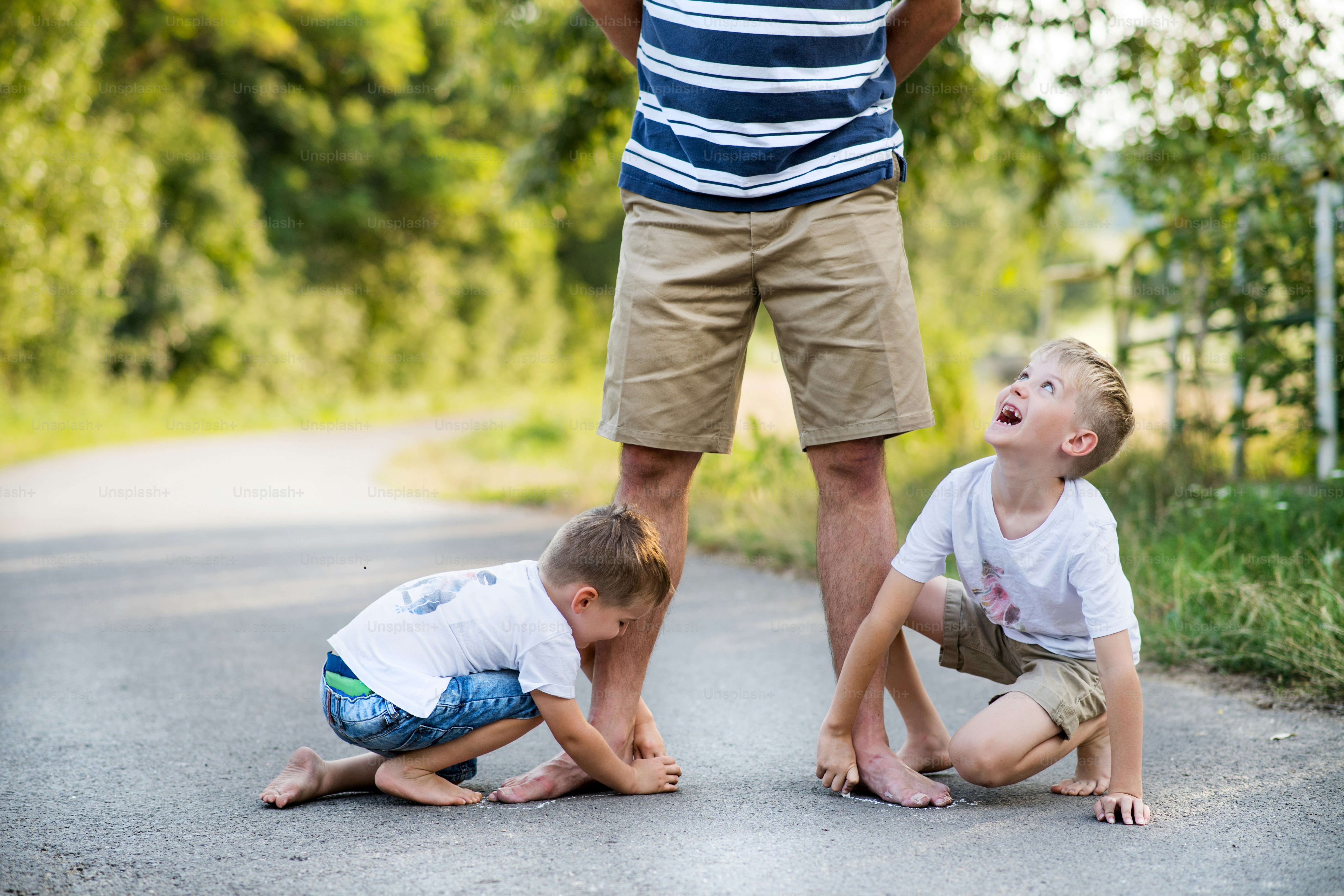 Two small sons playing with unrecognizable father on a road in park on a summer day, drawing with chalk