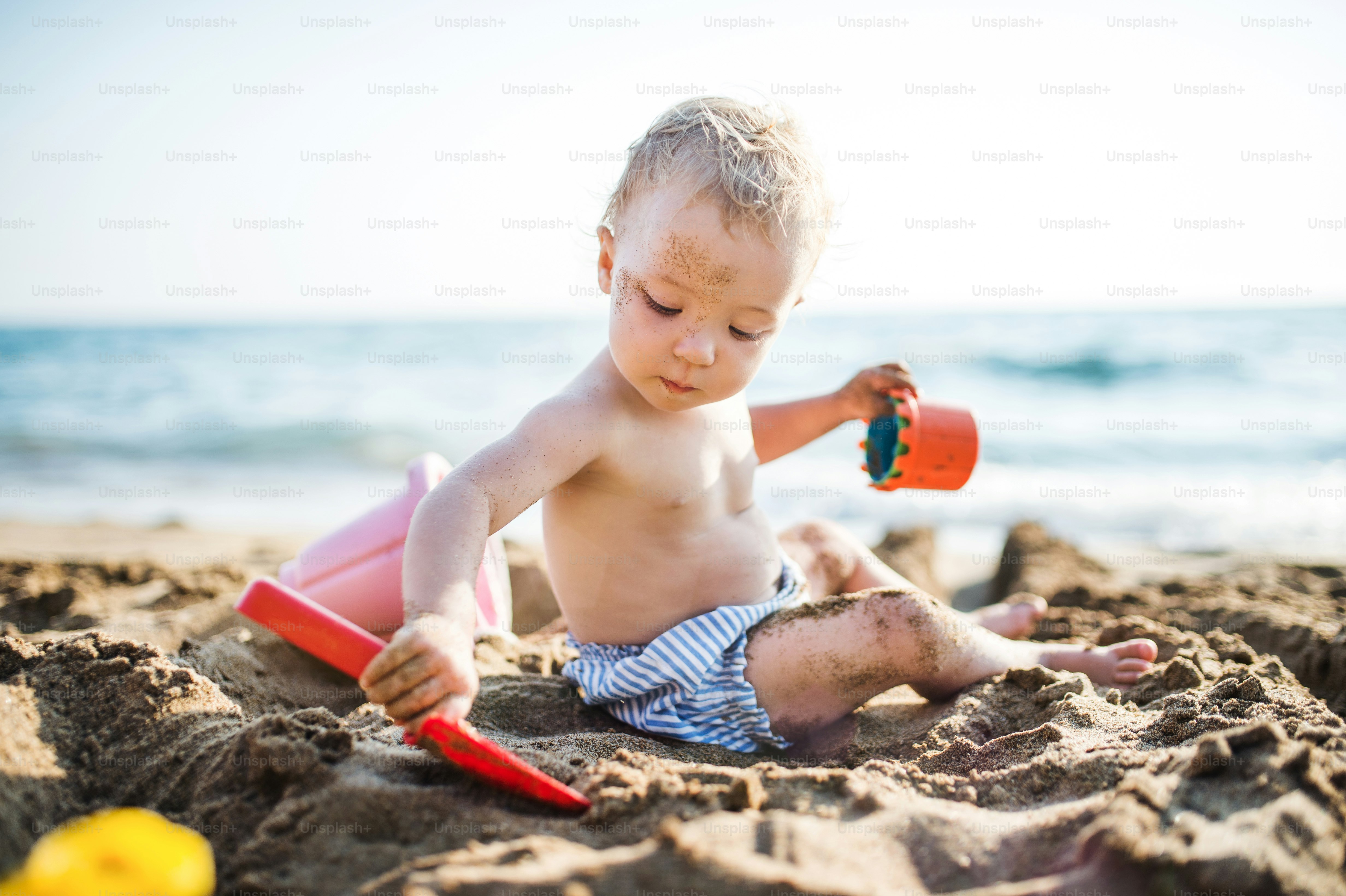 Ein kleines Oben-ohne-Kleinkind, das in den Sommerferien am Strand sitzt und im Sand spielt.
