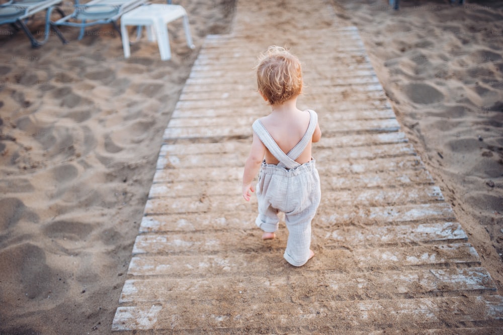 A rear view of small toddler girl walking topless on beach on summer holiday.