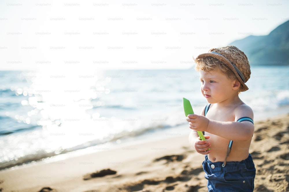 Un niño pequeño con sombrero y pantalones cortos parado en la playa en vacaciones de verano, sosteniendo una pala. Espacio de copia.