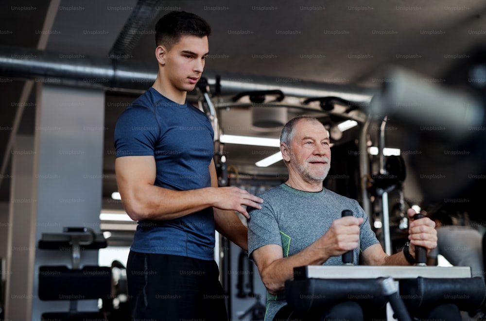 A senior man with a young male trainer doing strength workout exercise in gym.