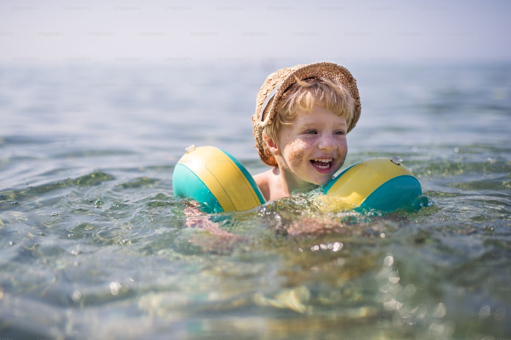 A happy small toddler boy with hat and armbands swimming in water on summer holiday, sand on cheeks.