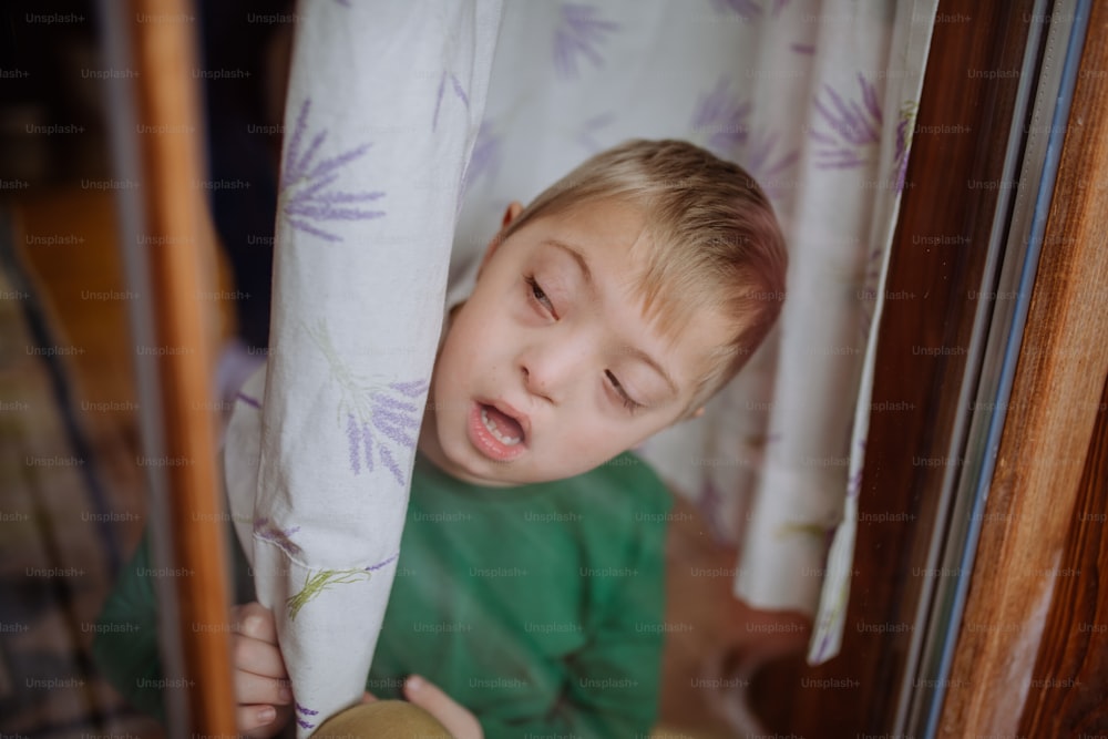 A little boy with Down syndrome looking through window at home.
