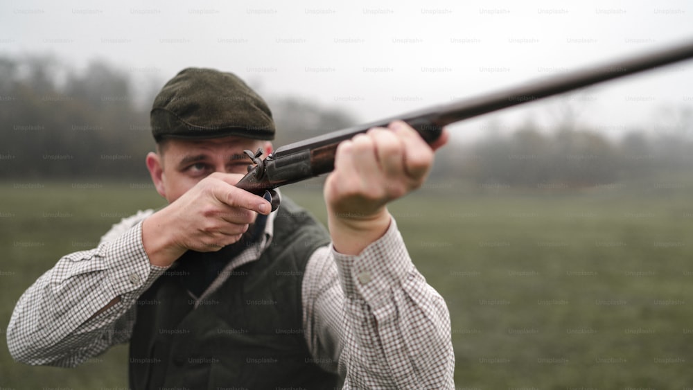 A hunter man in traditional shooting clothes on field aiming with shotgun.