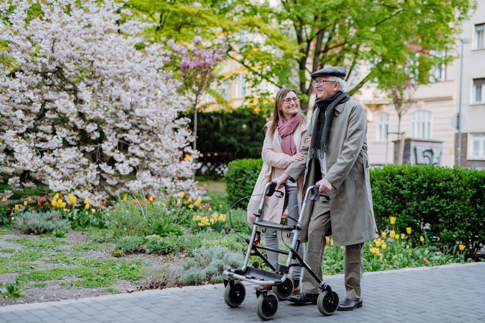 A senior man with walking frame and adult daughter outdoors on a walk in park.