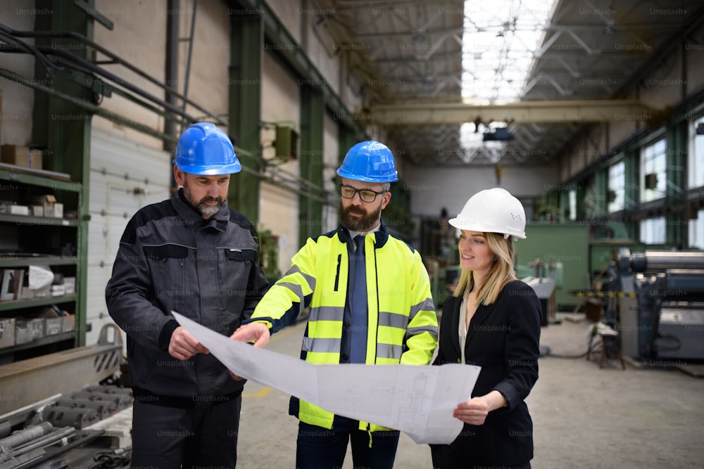 Um gerente supervisores, engenheiro e trabalhador industrial de uniforme discutindo plantas em grande salão de fábrica de metal.