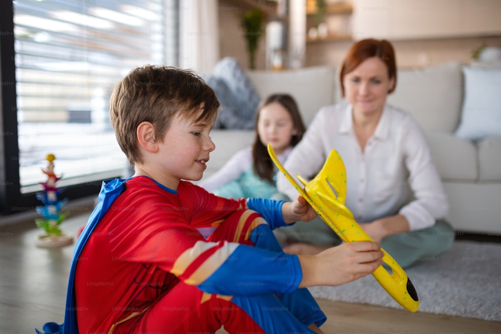 A little boy in superhero costume playing with aeroplane with his mother and sister at home.