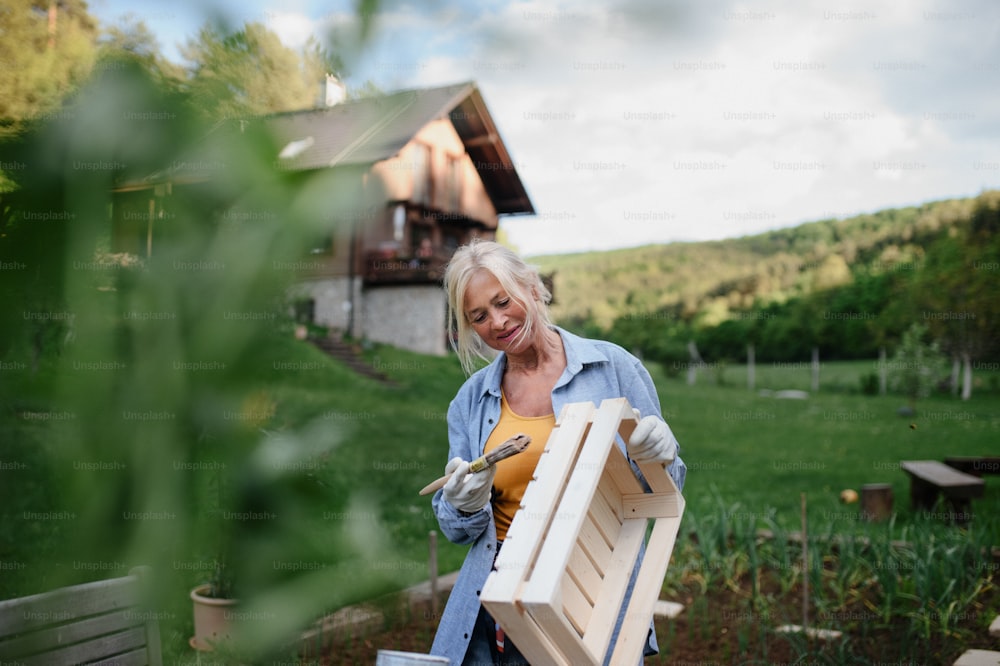 A senior woman with paintbrush impregnating wooden crate outdoors in garden.