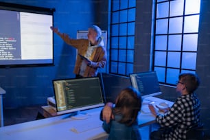Un profesor dando una conferencia de informática a jóvenes estudiantes en el cuarto oscuro. Cuarto. Explicación de la tecnología de la información.