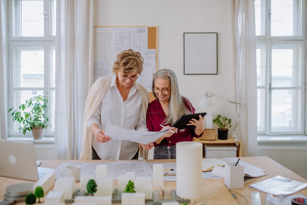 Mature women eco architects with model of modern bulidings and blueprints working together in an office.
