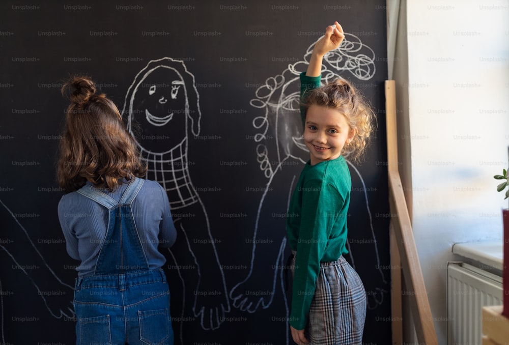 Two little girls drawing with chalks on a blackboard wall indoors in playroom, looking at camera.