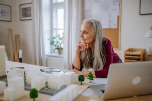 Une femme éco-architecte senior avec un modèle de bâtiments modernes travaillant dans un bureau.