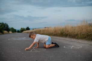 A little kid boy drawing with chalk on asphalt in summer.