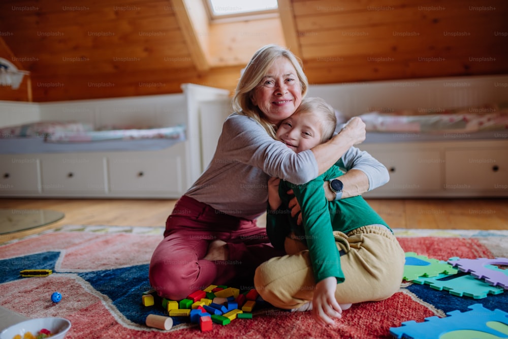 다운 증후군을 앓고있는 행복한 소년이 바닥에 앉아 집에서 할머니와 포옹하고 있습니다.